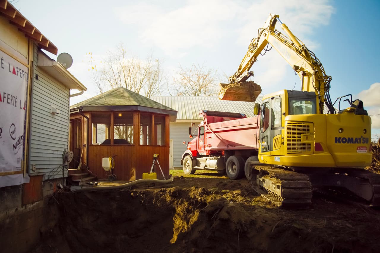 Excavatrice Komatsu creusant une fosse près d’une maison et d’un gazebo en bois, avec un camion benne rouge prêt à recevoir la terre.