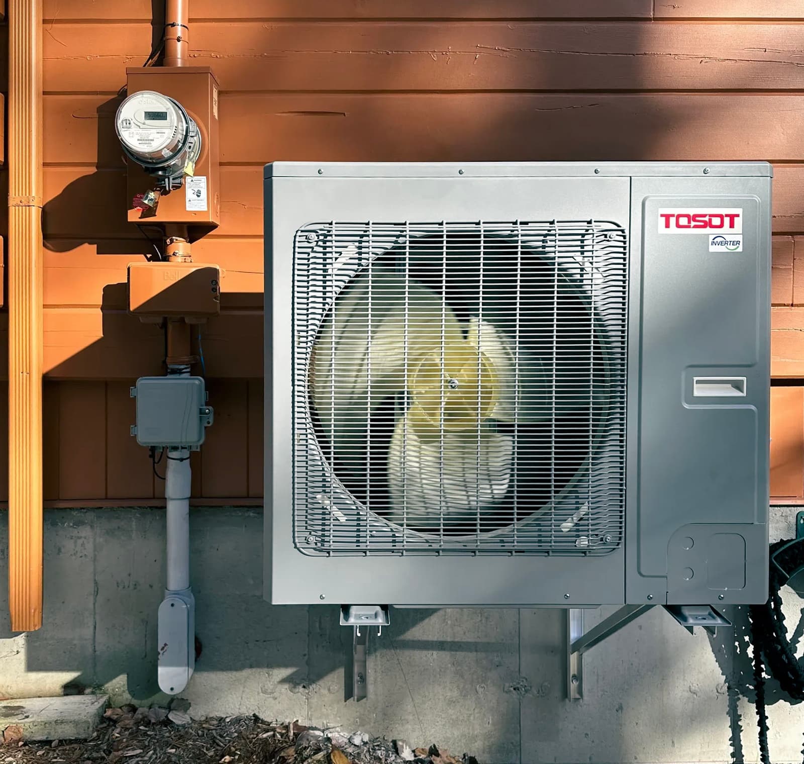 Unité extérieure d'une pompe à chaleur de la marque TOSOT installée sur le mur extérieur d'une maison, avec des conduits visibles et un compteur d'énergie à proximité, soulignant une solution moderne et efficace pour le chauffage et la climatisation.