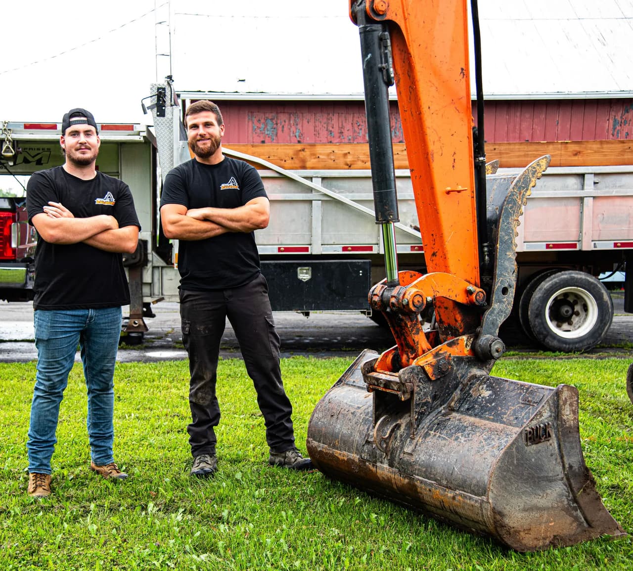 Deux ouvriers en vêtements de chantier posant fièrement devant une pelle mécanique orange et un camion de chantier sur un terrain gazonné