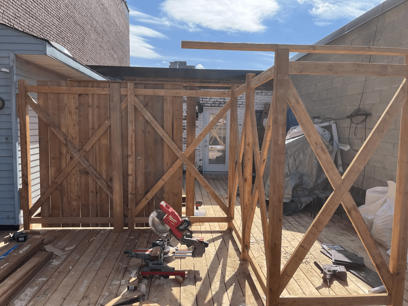Structure de clôture en bois en cours de construction sur une terrasse, avec panneaux verticaux, cadres renforcés en diagonale et outils de chantier visibles au sol.