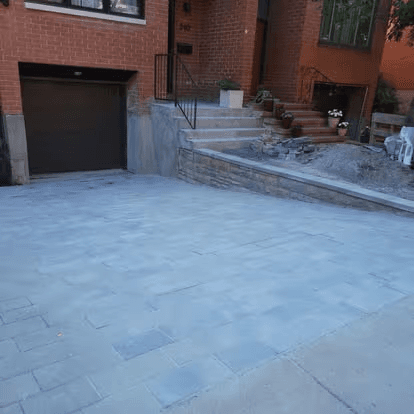 Entrée résidentielle en pavé uni gris devant une maison en brique, avec garage intégré, muret de soutènement et escalier extérieur en pierre