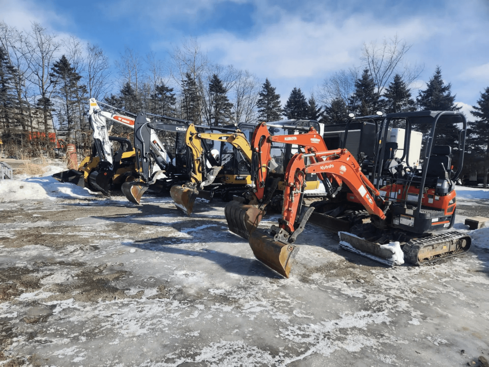 Alignement de mini-excavatrices et d’équipements de chantier sur chenilles stationnés sur un terrain enneigé, avec plusieurs modèles Kubota, Caterpillar, Bobcat et John Deere devant une zone boisée.