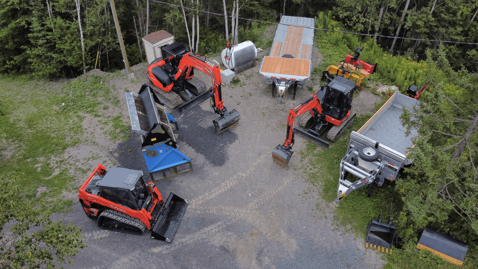 Vue aérienne d’un parc de machinerie lourde avec excavatrices compactes, chargeuse sur chenilles, remorques et accessoires d’excavation sur un terrain de gravier entouré de forêt.