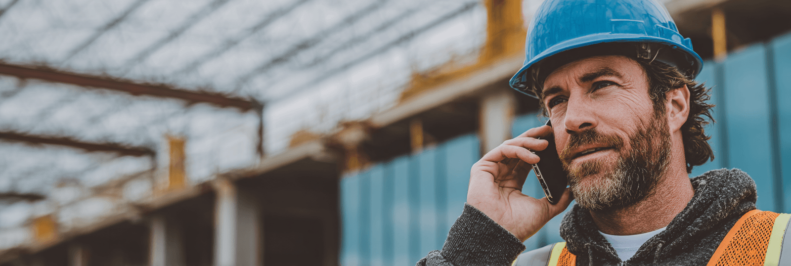 Professionnel de la construction portant un casque bleu et un gilet haute visibilité, parlant au téléphone sur un chantier de bâtiment en cours de construction
