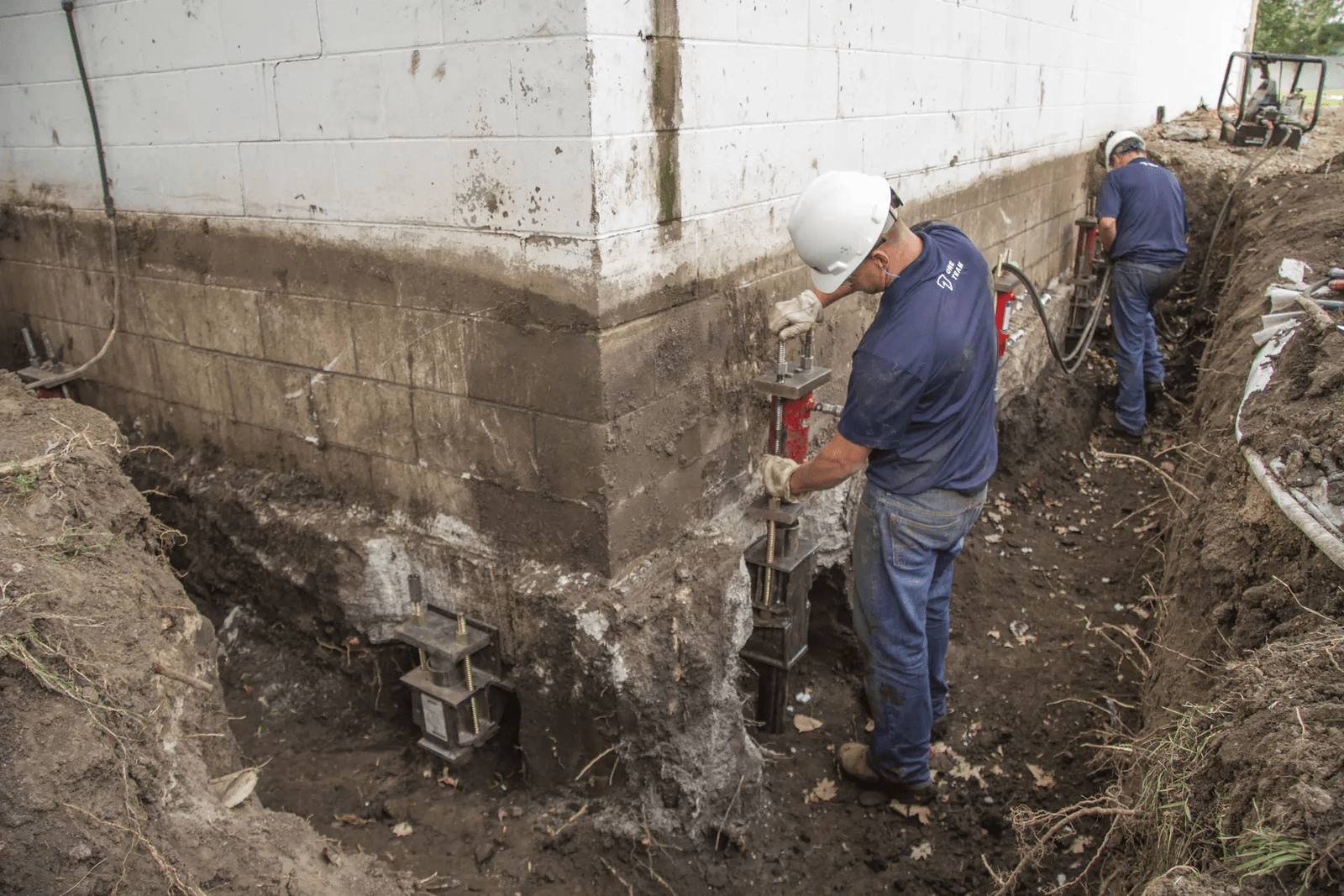 Travailleurs installant des supports métalliques de stabilisation le long d’une fondation en blocs de béton excavée lors de travaux de réparation structurale.