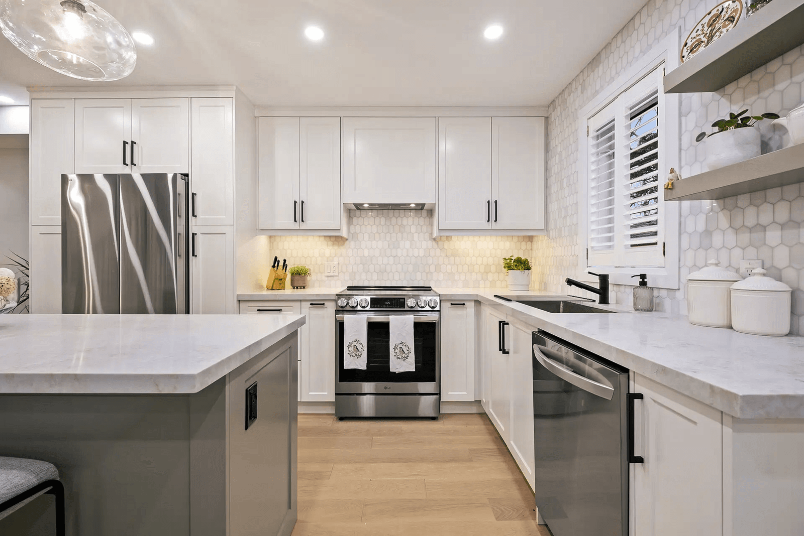 Cuisine moderne et lumineuse avec des placards blancs, un grand îlot central, des plans de travail clairs, une crédence en carrelage hexagonal, des appareils électroménagers en acier inoxydable et un évier noir.