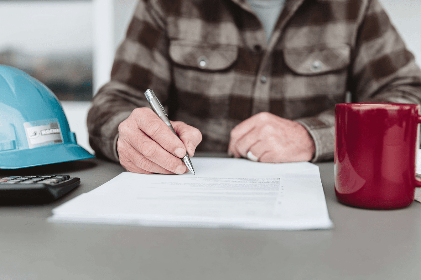 Entrepreneur en construction remplissant et signant des documents administratifs sur une table avec casque de chantier, calculatrice et tasse
