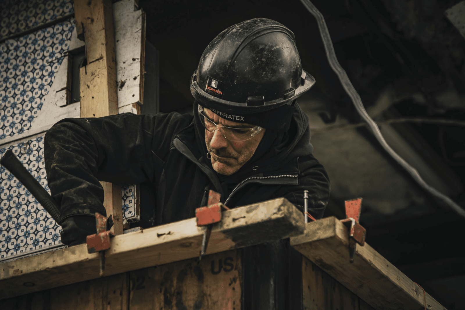Travailleur de la construction portant un casque et des lunettes de protection, effectuant des travaux de coffrage en bois sur un chantier de rénovation.