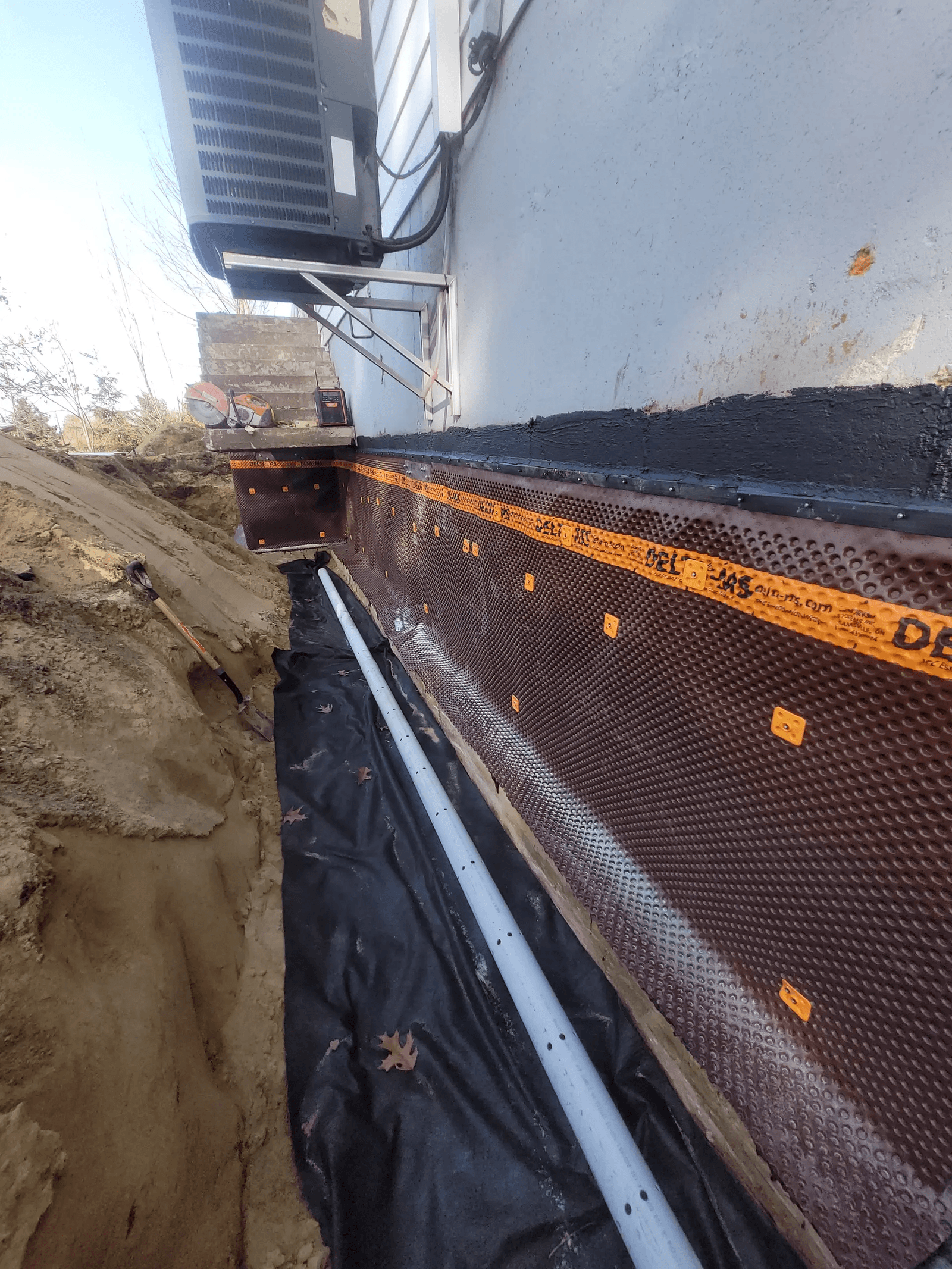 Drain français extérieur en installation le long d’une fondation avec membrane drainante, tuyau perforé et excavation autour de la maison