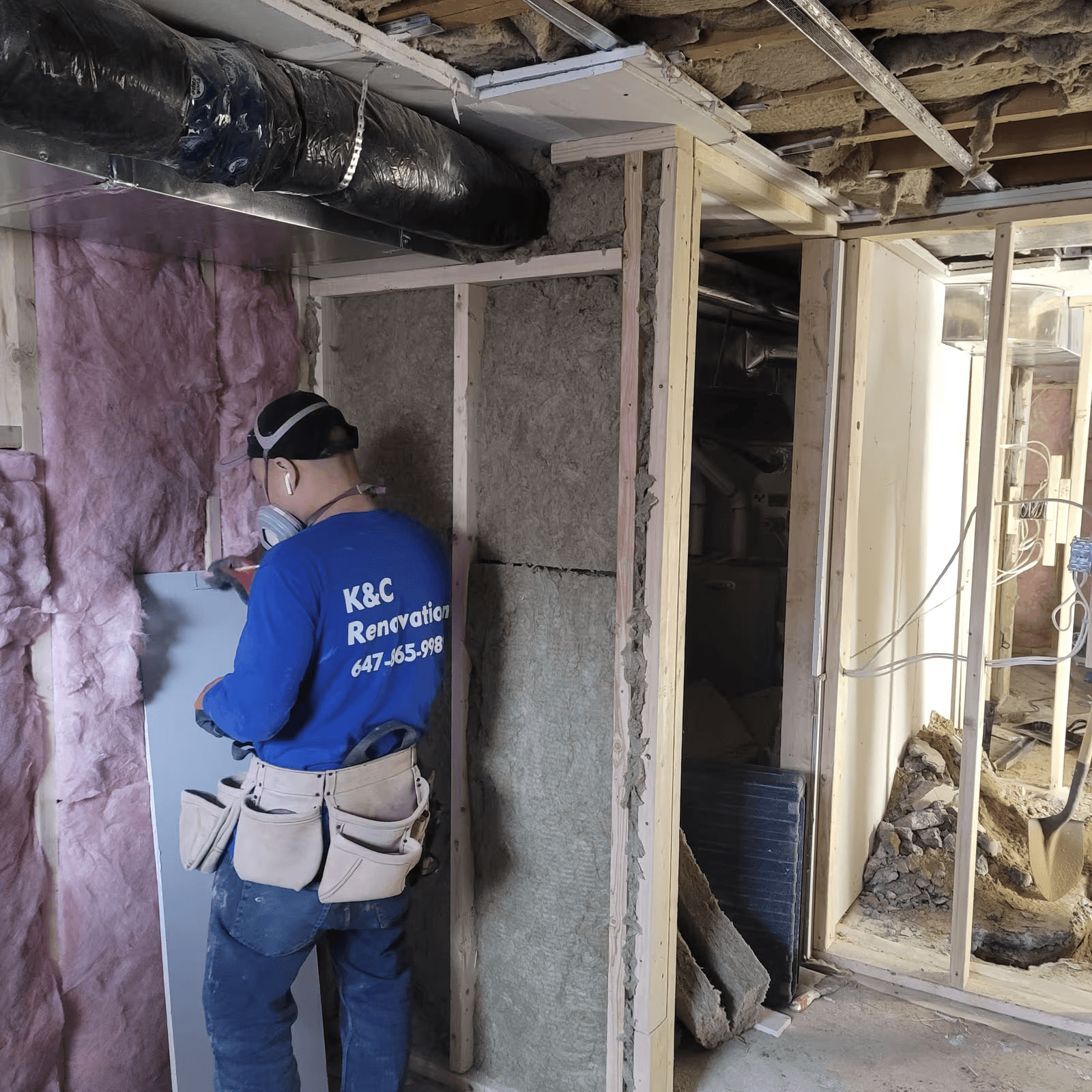 Travailleur en rénovation installant des panneaux de gypse dans une pièce en chantier avec ossature de bois, isolation en fibre de verre et conduits de ventilation apparents.