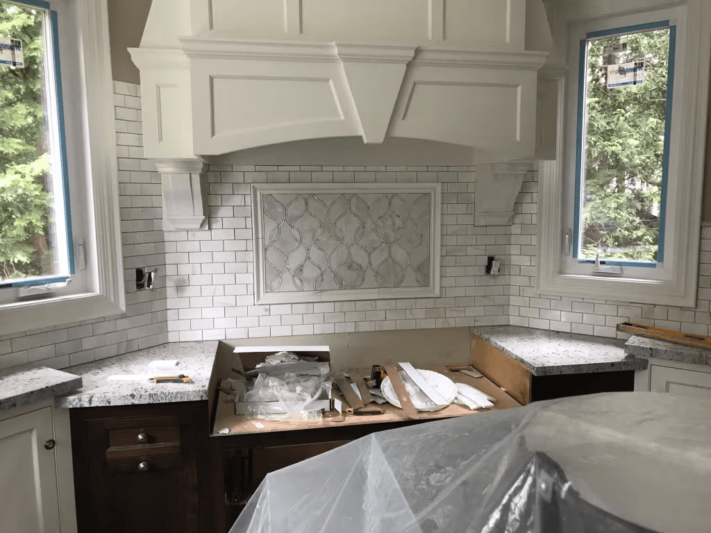 Cuisine résidentielle en rénovation avec armoires sur mesure, dosseret en carreaux blancs, comptoirs de pierre et hotte décorative en cours de finition