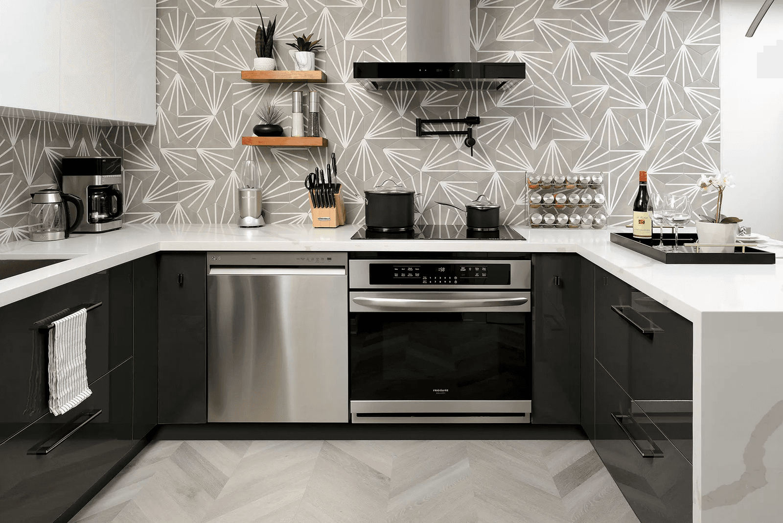 Cuisine contemporaine avec des placards noirs, des plans de travail blancs, une crédence aux motifs géométriques, des appareils électroménagers en inox, une hotte noire et un sol à motif chevrons.