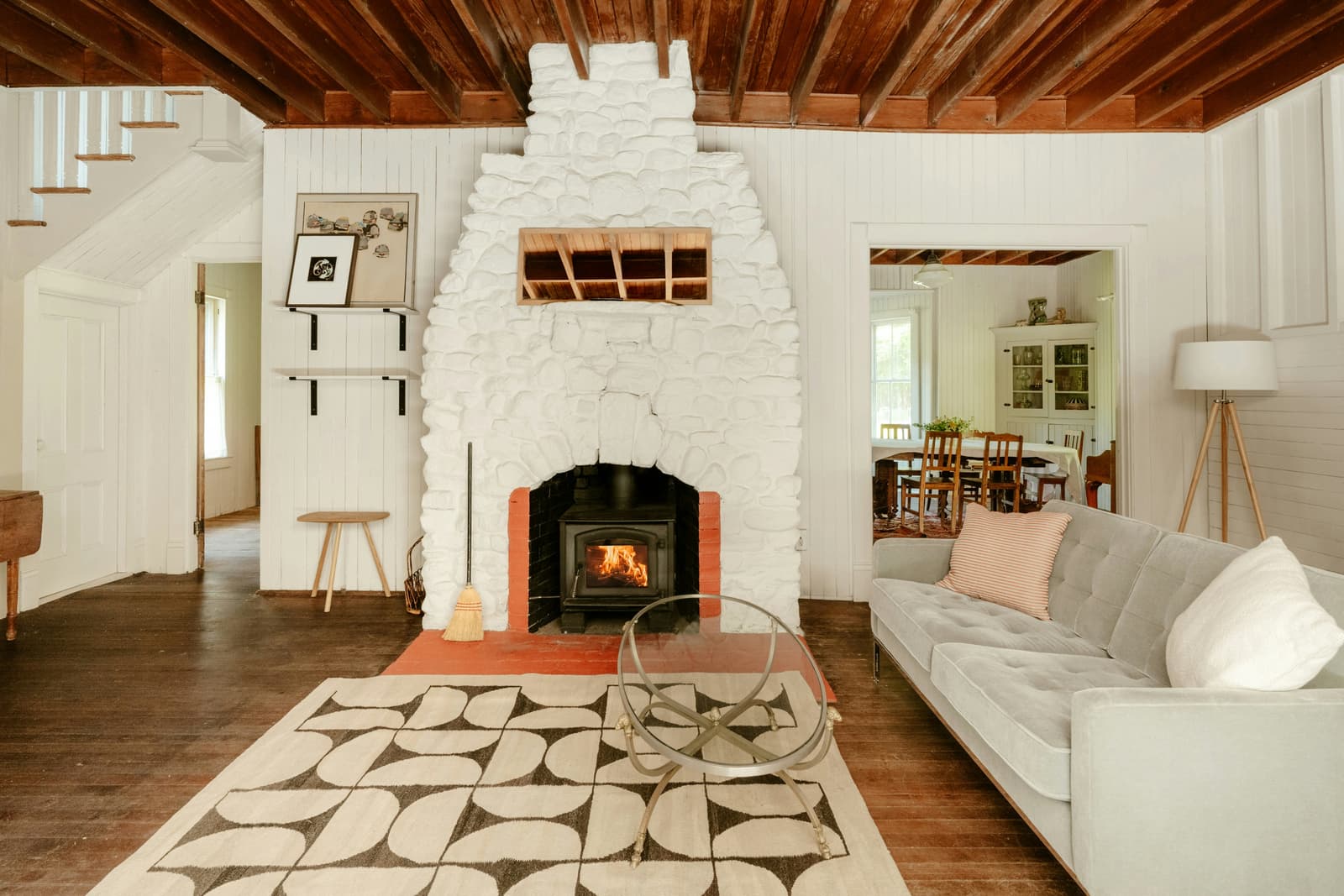 Salon lumineux au style rustique avec foyer en pierre blanche, poêle à bois allumé, plafond à poutres apparentes, plancher en bois et mobilier contemporain.