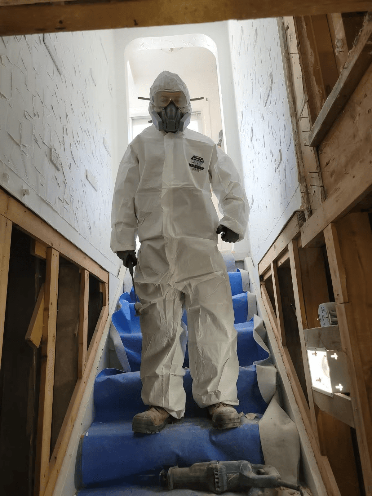  Travailleur en combinaison de protection avec respirateur sur un escalier en rénovation, entouré de murs ouverts et de charpente apparente / Worker wearing a protective suit and respirator on a staircase under renovation, surrounded by open walls and exposed framing 