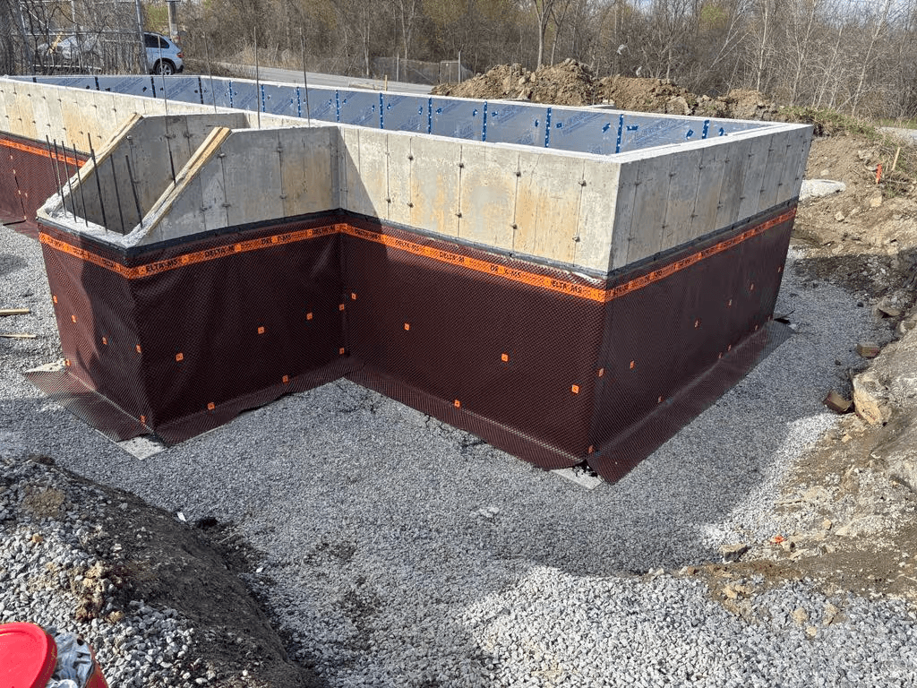  Fondation résidentielle en béton avec membrane drainante et imperméabilisante installée autour des murs, sur lit de gravier avant remblaiement