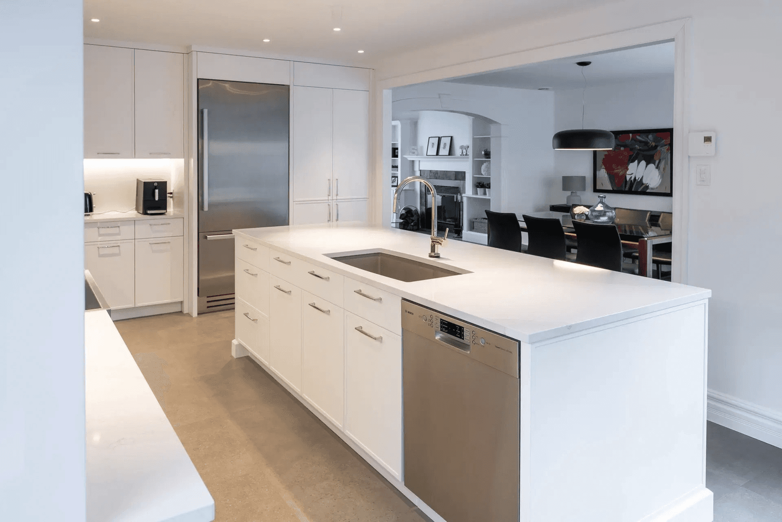 Cuisine blanche moderne avec grand îlot central, évier intégré, électroménager en acier inoxydable, placards toute hauteur et ouverture sur une salle à manger contemporaine.