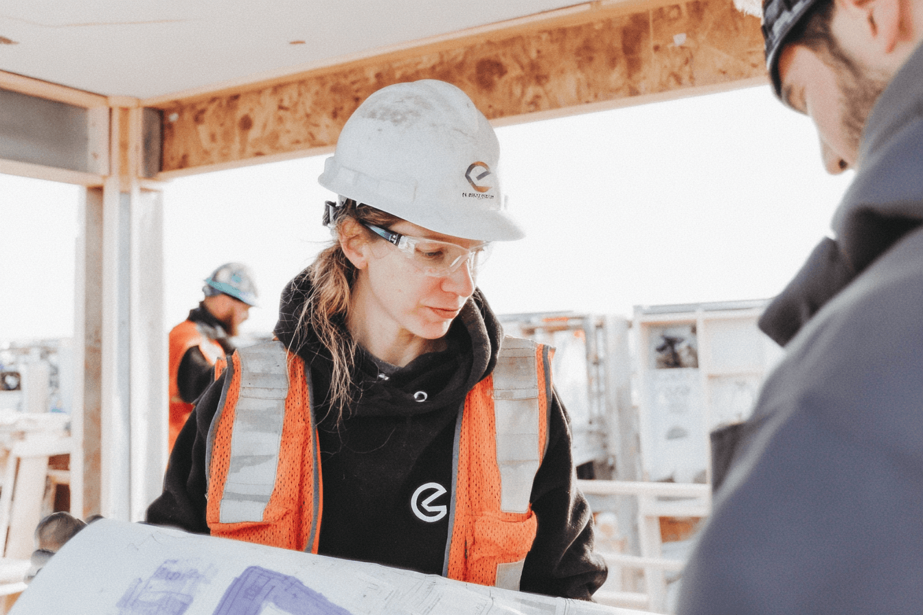 Professionnelle de la construction portant un casque blanc, des lunettes de sécurité et un gilet haute visibilité, examinant des plans sur un chantier intérieur en rénovation