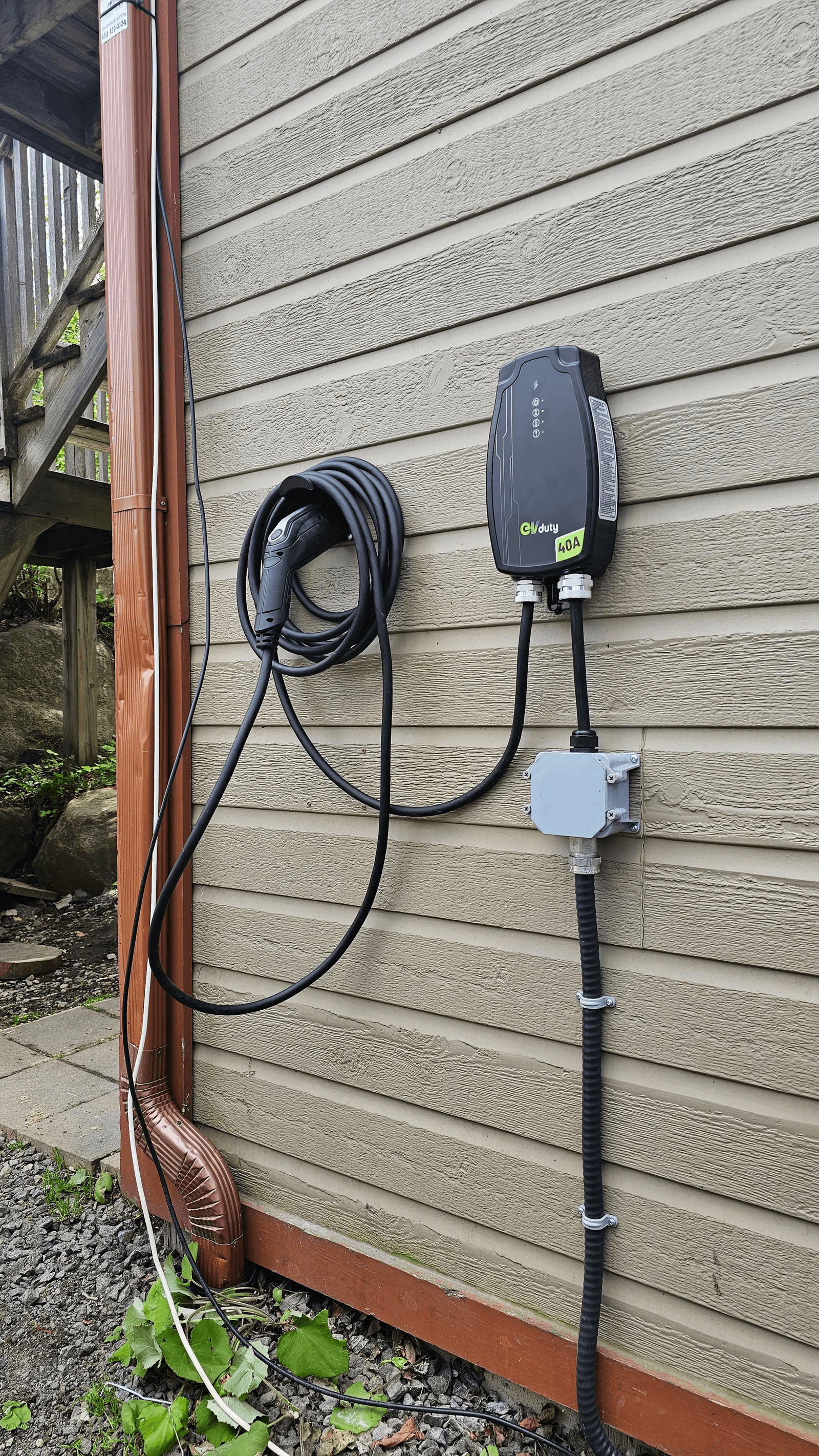 Borne de recharge électrique EVduty de 40 ampères installée sur le revêtement extérieur d'une maison, avec câble de recharge enroulé, connecteur et raccordement électrique apparent / EVduty 40-amp electric vehicle charging station installed on a house's exterior siding, featuring a coiled charging cable, connector and exposed electrical connections