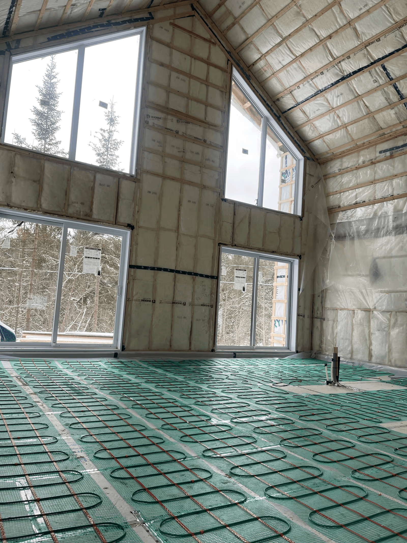 Grande pièce résidentielle en construction avec plancher chauffant radiant installé, murs et plafond isolés, grandes fenêtres et préparation pour finition intérieure
