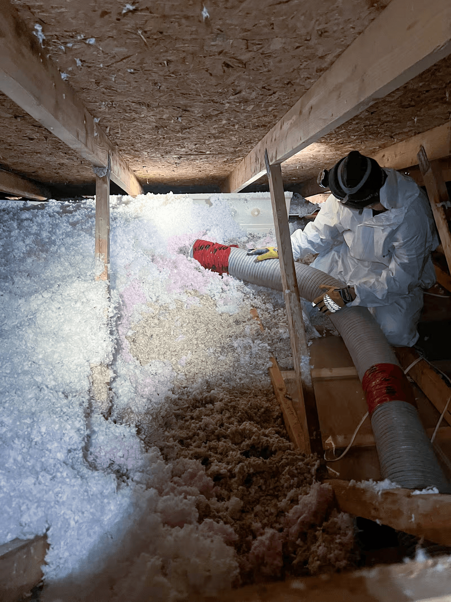 Technicien en isolation travaillant dans un grenier avec équipement de protection, tuyau de soufflage et isolant en vrac installé entre les éléments de charpente.