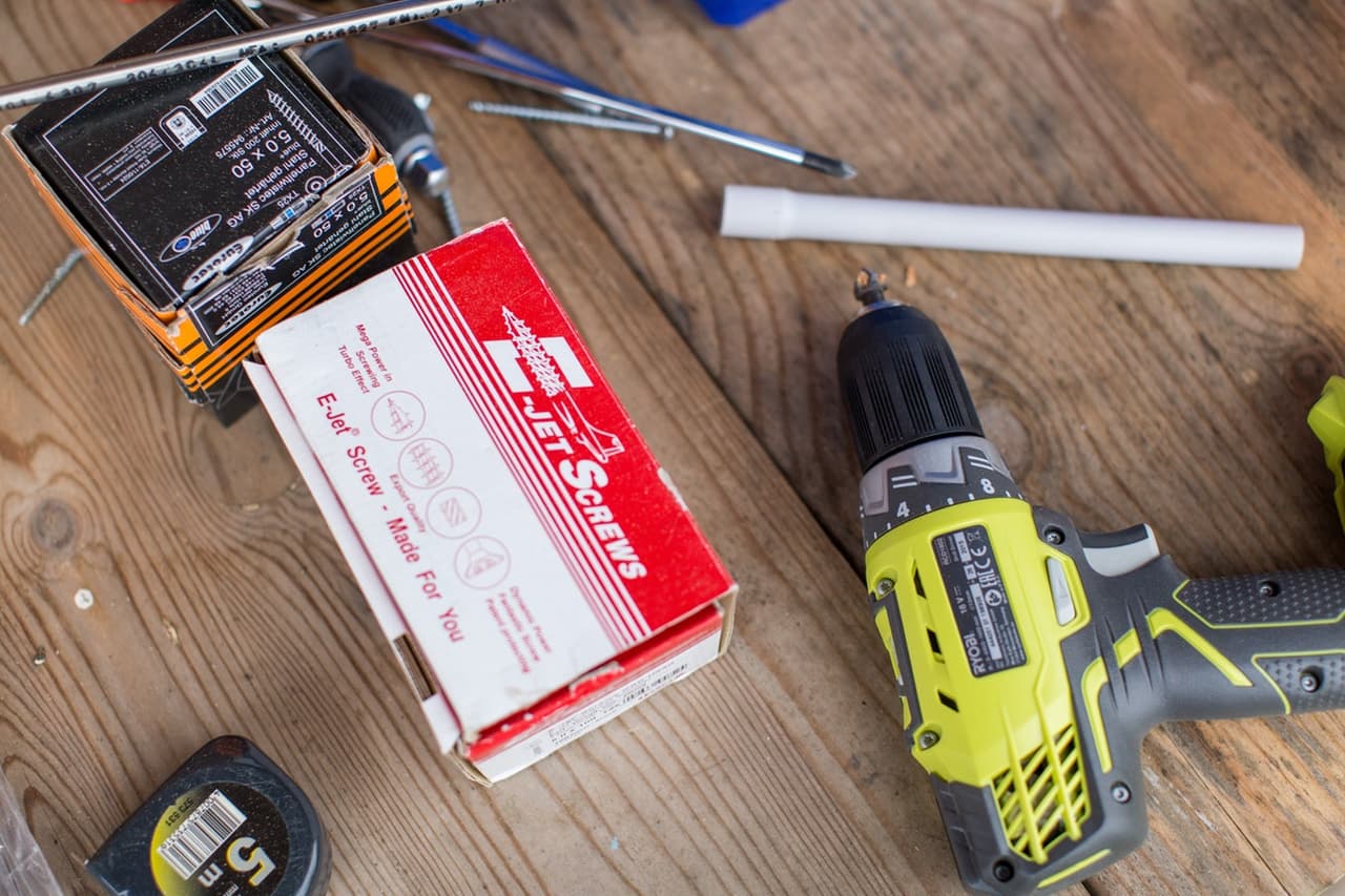Vue de haut d'outils de rénovation posés sur une table en bois, incluant une perceuse jaune et noire, un ruban à mesurer et des boîtes de vis de marque Jet Screws