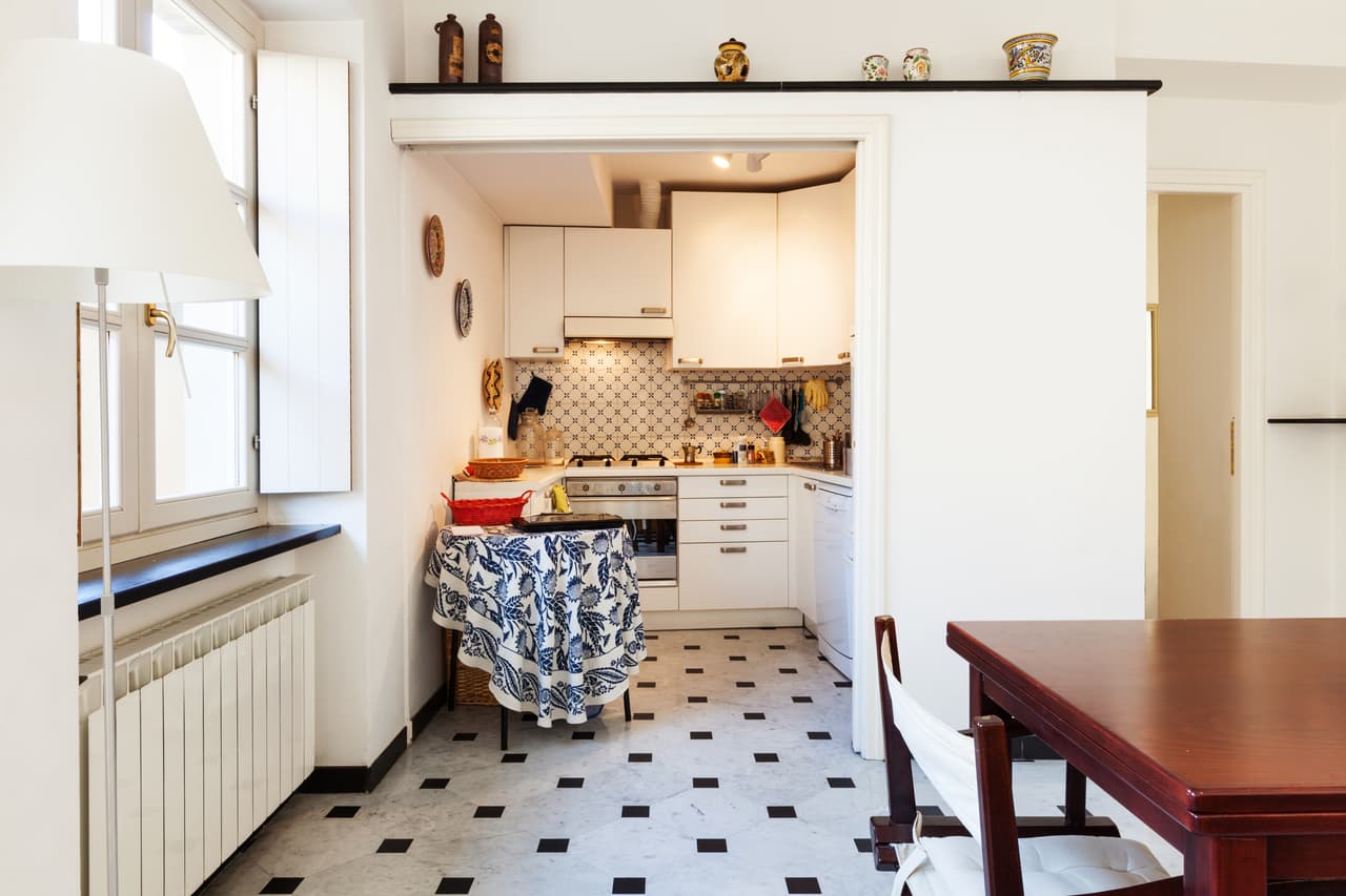 Intérieur résidentiel montrant une table de salle à manger au premier plan et une ouverture menant à une cuisine blanche avec un sol en carreaux de damier noir et blanc